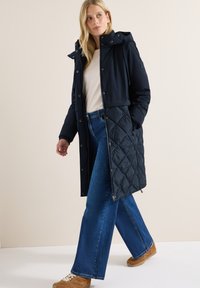 Cappotto imbottito blu navy con motivo trapuntato, cappuccio e bottoni a pressione, abbinato a jeans blu a gamba larga e sneakers beige.