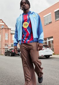 Casaco azul com fecho éclair em cima de uma camisa listrada em vermelho e azul com um logótipo amarelo em forma de círculo. Calças largas xadrez em tons de castanho. Ténis completam o visual.