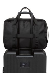 Musta pyörällinen matkalaukku, jossa on teksturoitu kova kuori ja lyhyempi musta kangaskassi päällä, varustettuna kaksilla kahvoilla ja vetoketjullisella taskulla.