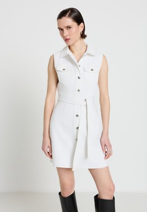 Frau trägt ärmelloses weißes Kleid mit Knopfleiste, Kragen und Gürtel, kombiniert mit schwarzen kniehohen Stiefeln, steht vor einfachem Hintergrund.