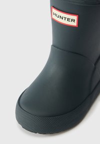 Bota de borracha preta com uma sola texturizada. Apresenta um rótulo vermelho e creme com "HUNTER." Ponteira arredondada e design superior liso.