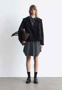 Black double-breasted wool-blend coat with wide lapels, worn over a black and white striped shirt dress. Brown suede bag and black shoes with socks.