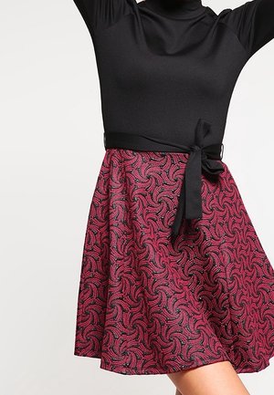 Persoon draagt een zwarte top met lange mouwen en een hoge hals, en een roze patroon rok die met een zwarte sjerp in de taille is gestrikt.