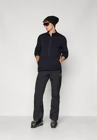 Pile nero con cerniera frontale, collo alto e maniche lunghe, abbinato a pantaloni da neve neri e scarponi da sci. Indossato con un berretto e occhiali da sole.