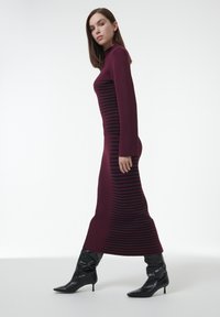 Bordeaux gebreide jurk met lange mouwen en horizontale zwarte strepen; gedragen met zwarte kniehoge laarzen en een getailleerd silhouet.