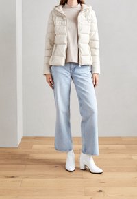 Lys beige puffjakke med hætte, lynlåslommer, beige top, lyseblå brede bukser og hvide ankelstøvler på et trægulv.