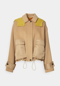 Veste en laine beige avec poches avant oversize, renforts jaunes aux épaules, poignets ajustables, col, fermeture par boutons et ourlet à cordon de serrage.