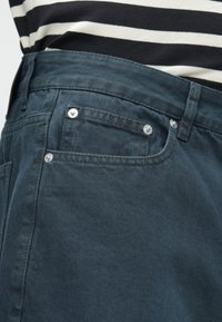 Jeans de ganga escuro com rebites metálicos nos bolsos da frente, uma textura suave e uma cintura média, complementados por uma camisa riscada por cima.