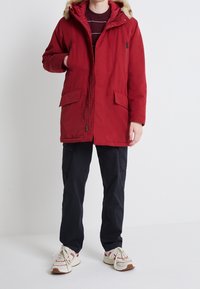 Röd parkas med foder av konstpäls i huvan, dragkedja fram, två stora fickor fram och en avslappnad passform. Bärs över mörka cargobyxor och sneakers.