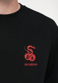 T-shirt nera in cotone con grafica di un serpente rosso e il testo "HAN KJØBENHAVN" sotto. Collo arrotondato, maniche corte, texture liscia.