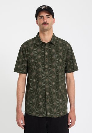 Mann mit Schnurrbart, der eine schwarze Kappe und ein grün gemustertes kurzärmeliges Hemd mit Knöpfen trägt, steht vor einem einfarbigen Hintergrund.