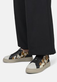 Sneaker in pelle e tessuto con un motivo maculato marrone e bianco, accenti neri e una suola beige testurizzata. Il design presenta lacci e un profilo basso.