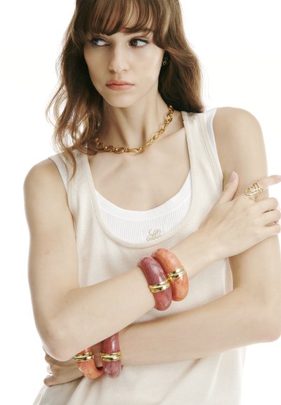 Fiatal nő világosbarna hajjal, réteges ujjatlan felsőket visel, nagy rózsaszín és korallszínű karkötőkkel, aranylánccal és arany gyűrűvel, oldalra néz.