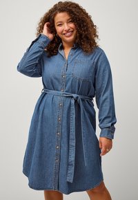 Abito camicia in denim con maniche lunghe, chiusura con bottoni, taschino sul petto e cintura rimovibile. Presenta un orlo curvo e un colore blu medio.