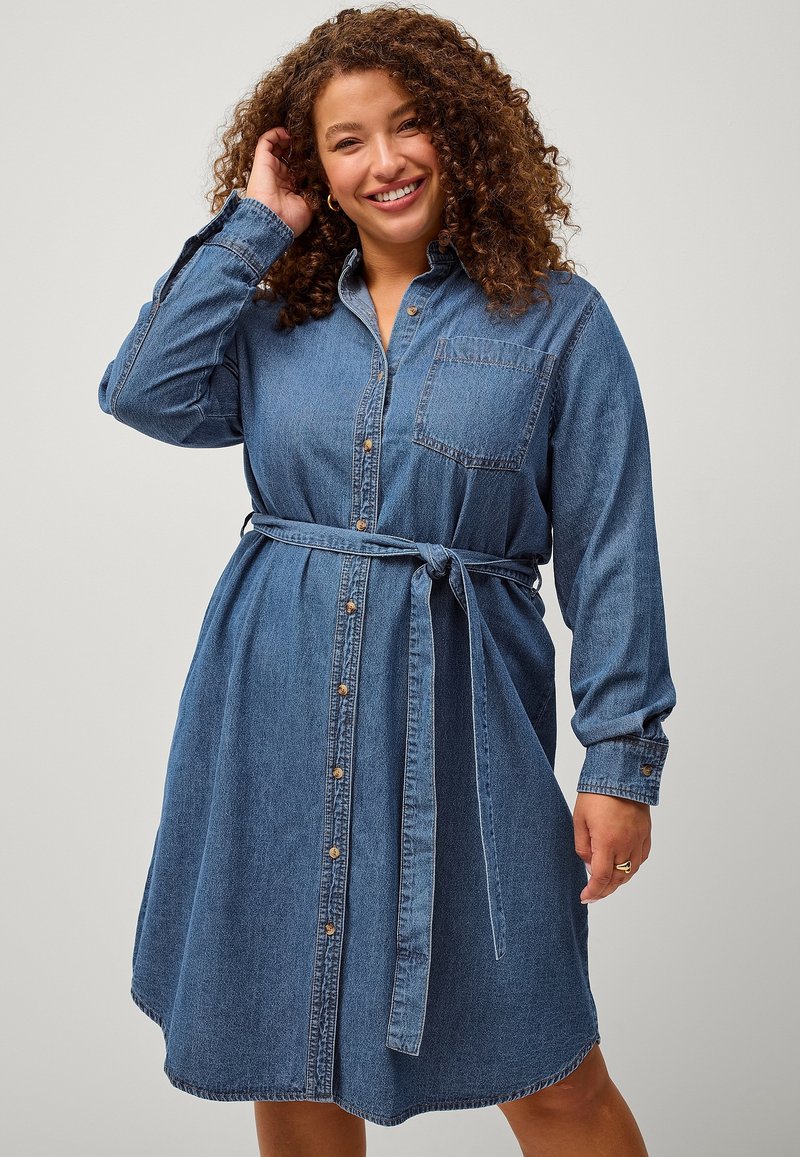 Abito camicia in denim con maniche lunghe, chiusura con bottoni, taschino sul petto e cintura rimovibile. Presenta un orlo curvo e un colore blu medio.