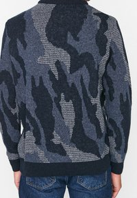Person som bär en mörkblå och grå camouflage-mönstrad tröja och blå jeans, visad från ryggen mot en enfärgad bakgrund.