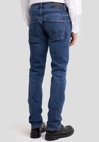 Egy férfi, kék farmernadrágot visel, hátul zsebekkel, fekete bőrcipővel és fehér hosszú ujjú inggel, háttal áll.