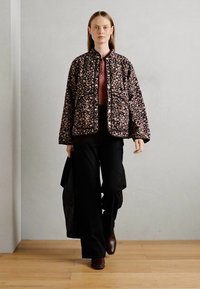 Woman walking indoors wearing a floral quilted jacket, pink shirt, black wide-leg pants, brown shoes, and carrying a black bag.