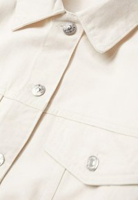 Beige cotton shirt featuring silver snap buttons, a collar, and two chest pockets with flaps. The texture is smooth with a slightly ribbed finish.