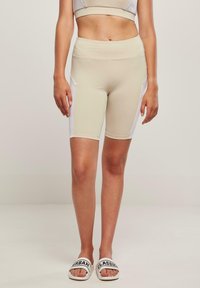 Bézs kerékpáros nadrág fehér oldalsó panelekkel, magas derékkal és rugalmas anyagból. A modell fehér szandált visel fekete szöveges díszítéssel.