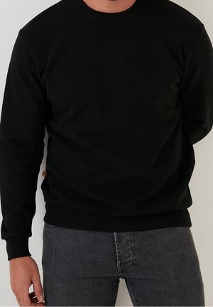 Uomo con maglione nero semplice a girocollo e jeans grigio scuro, in piedi contro un muro chiaro con le mani dietro la schiena.