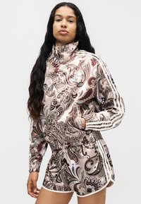 Femme portant un ensemble veste et short assortis à motif paisley, col zippé et bandes blanches sur les côtés, debout avec une main dans la poche.