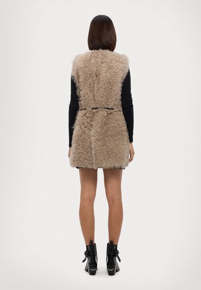 Beige nep-bont vest met een fitted silhouet, vastgebonden in de taille. Draag het over een zwart langmouwen top, gecombineerd met zwarte enkellaarsjes.