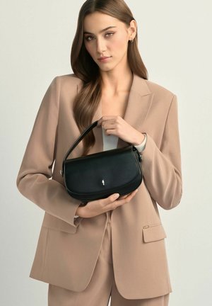 Młoda kobieta w beżowym garniturze trzymająca małą czarną skórzaną torebkę z jedną górną rączką na jednolitym tle.