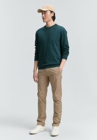Sudadera verde oscura, chinos beige y zapatillas blancas; gorra también beige; ajuste informal y textura resaltada, con líneas simples y sin patrones.
