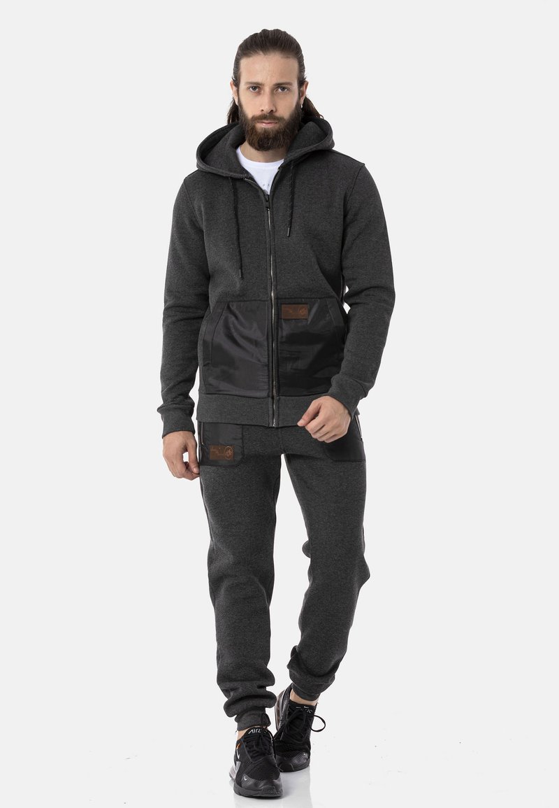 Gray zip-up hoodie with a black panel and two front pockets, paired with matching joggers. Textured fabric, adjustable hood, and logo accents.