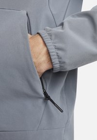 Grijze jack met een ritszak, gladde textuur, elastische manchetten en een subtiele glans. Voorzien van een rits met een stoffen lus.