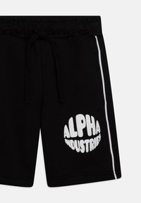 Melnas šortos, izgatavotas no mīksta auduma, ar aukliņu jostu, baltu sānu joslu un lielu baltu "ALPHA INDUSTRIES" logo.