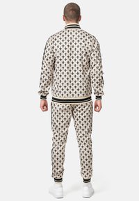 Beige tracksuit with black fleur-de-lis pattern, featuring a striped collar and cuffs, ribbed waist and cuffs, and white sneakers.