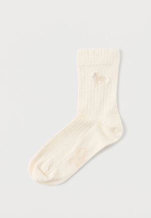 Șosetă crem cu nervuri și un mic detaliu brodat cu un cal lângă marginea superioară, așezată plat pe o suprafață albă.