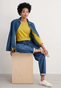 Denimist jope, kollase pikkade varrukatega särgi peal, helesinisest dressipükstest ja valgetest tossudest koosnevas riietuses. Puitplokk istumise asemel.