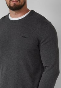 Grijs gebreid sweater met een ronde hals. Heeft een klein zwart logo op de borst. De stof lijkt zacht en textuur te hebben.