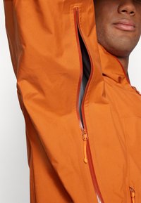 Veste imperméable orange avec une texture lisse, dotée d'une ventilation zippée sur la manche gauche et d'un zip contrastant rouge.