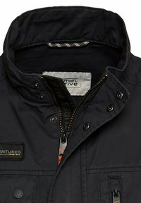 Marineblaue Jacke mit hohem Kragen, Reißverschluss vorne, Druckknöpfen und einem schwarzen Etikett. Verfügt über einen strukturierten Stoff und orangefarbene Akzente.