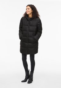 Crni dugi kaput s podstavom, patentnim zatvaračem sprijeda, kapuljačom i bočnim džepovima, uparen s crnim uskim trapericama i crnim gležnjačama na visoku petu.