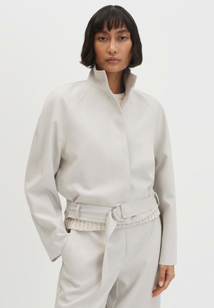 Vrouw met kort zwart haar, gekleed in een lichtgrijze jas met riem over een crèmekleurige gebreide trui, staand tegen een eenvoudige lichte achtergrond.