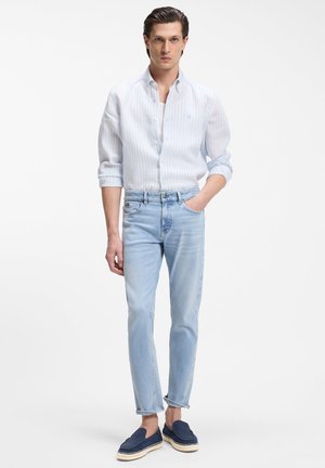 Man draagt lichtblauw gestreept overhemd met knopen, ingestopt in lichtblauwe spijkerbroek, met één hand in de zak en donkerblauwe instapschoenen.