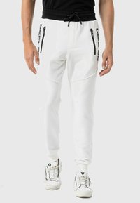 Weiße Sweatpants mit einem Kordelzugbund, seitlichen Reißverschlusstaschen und schwarzen Markenzeichen entlang des linken Beins. Glatter, dehnbarer Stoff.