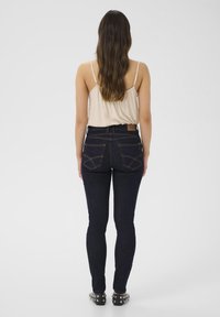 Tumši zili pieguļoši džinsi ar gludu tekstūru un redzamām šuvju detaļām, valkāti ar gaiši beidzīgu bez piedurknēm kretu, skatīti no aizmugures.