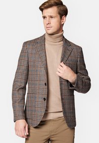 Brązowa marynarka w kratę z niebieskimi akcentami, szpiczaste klapy, dwa guziki i teksturowana tkanina, noszona na beżowym swetrze z wysokim kołnierzem.