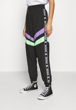 Person wearing black track pants with purple and green stripes and KANI logo stripes on the sides, paired with black high-top sneakers.