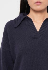 Femme portant un pull en maille côtelée bleu marine foncé avec un large col en V et un col plié, dévoilant son cou et ses lèvres.