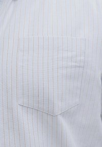 Λευκό ύφασμα με λεπτές κάθετες καφέ λεπτομέρειες και ένα τετράγωνο τσεπάκι στο στήθος ραμμένο στο μπροστινό μέρος.