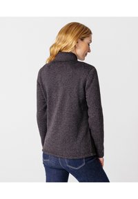 Chaqueta de forro polar gris oscuro con cuello alto, tejido texturizado y dobladillo curvado, combinada con jeans azules. Sin hardware ni patrones visibles.
