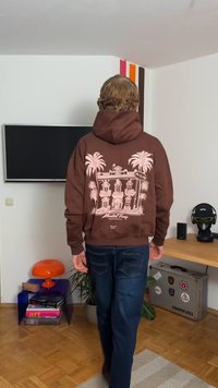 Καφέ φούτερ με γραφικό σχέδιο που απεικονίζει φοίνικες και ένα κτίριο στο πίσω μέρος. Το υλικό φαίνεται απαλό και περιλαμβάνει κουκούλα με κορδόνι.