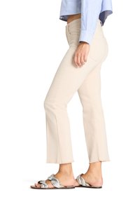 Mujer con pantalones cortos beige, camisa de manga larga azul claro y sandalias planas trenzadas plateadas, de pie de perfil.